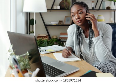 Serious young Black woman in loungewear talking on phone with colleague, reading document on laptop screen and taking notes