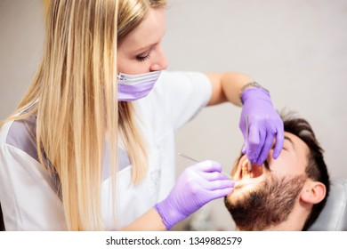 Serious Young Blond Haired Female Dentist Examining Young Male Patient In Dental Clinic