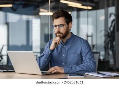 Serious young business man works in the office with a laptop, sits at the desk and looks thoughtfully at the screen, holding his chin with his hand. - Powered by Shutterstock