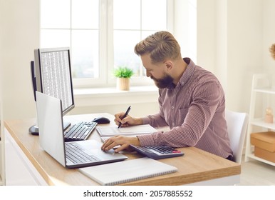 Serious Young Businessman Or Financial Accountant Sitting In Office Workplace At Desk With Desktop And Laptop Computers, Writing Something, Doing Multiple Tasks, Working With Data Files, Taking Notes