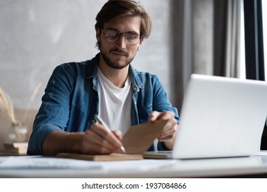 Serious young businessman with the glasses sitting and using laptop at home office and taking notes. - Powered by Shutterstock