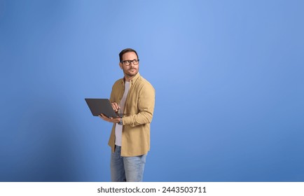 Serious young businessman holding laptop and looking away thoughtfully over isolated blue background - Powered by Shutterstock