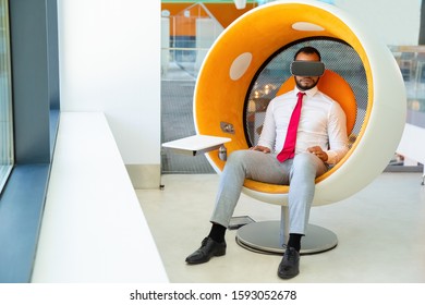 Serious young businessman using VR headset. Full length view of young African American businessman sitting in spherical chair and using virtual reality headset. Technology concept