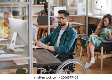 Serious young businessman in wheelchair sitting at the table and typing on computer he working at office with his colleagues - Powered by Shutterstock