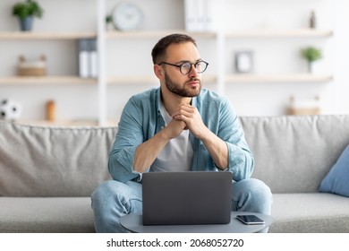 Serious Young Caucasian Guy Sitting On Couch In Front Of Laptop Computer At Home. Thoughtful Millennial Man Daydreaming While Working Online, Using Portable Pc Indoors