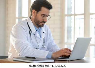Serious young Caucasian male doctor in white medical uniform sit at desk in hospital consult client on laptop online, man GP or physician fill patient medical card use Web application form on computer