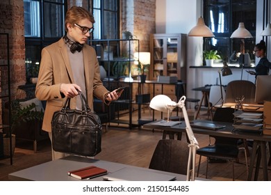 Serious Young Caucasian Man In Eyeglasses Holding Bag And Checking Taxi App On Phone While Leaving Office At Night