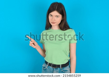 Serious Young caucasian woman wearing green T-shirt over blue background smirks face points away on copy space shows something unpleasant. Look at this advertisement. Big price concept