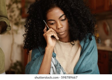 Serious young dark-skinned female with Afro hairstyle having worried and unhappy look while talking on mobile phone, receiving bad negative news, sitting alone at kitchen table wearing wrap - Powered by Shutterstock