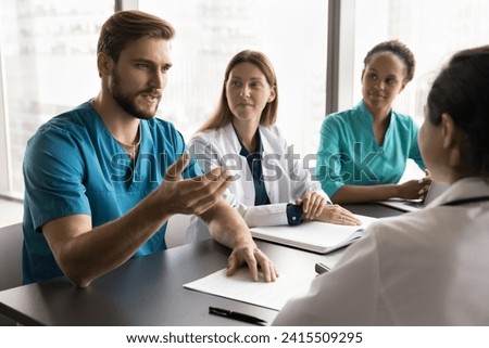 Serious young doctor man speaking to diverse colleagues, discussing job, medical profession, giving surgeon expertise, consultation for difficult patient case. Clinic staff talking at meeting table