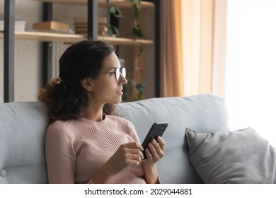 Serious young dreamy woman in eyeglasses sit on sofa in living room holding in hands smartphone looking to aside dreaming or thinking. Leisure and modern tech, new mobile app, wait for call concept