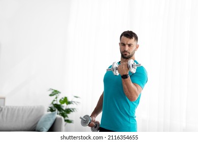 Serious young european guy doing strength exercises for arms, lifting dumbbells in room interior, on window background. Gym at home and new normal during covid-19 quarantine, muscle workout alone