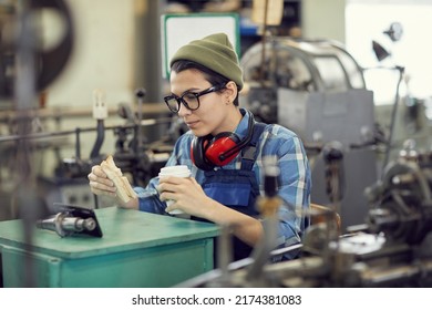 Serious young factory employee in hipster hat and glasses sitting at factory workshop and watching video on smartphone leaned on metal detail while eating sandwich - Powered by Shutterstock