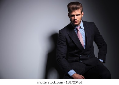 Serious Young Fashion Business Man In Classic Suit And Tie Is Sitting On A Chair And Looking At The Camera On Gray Background