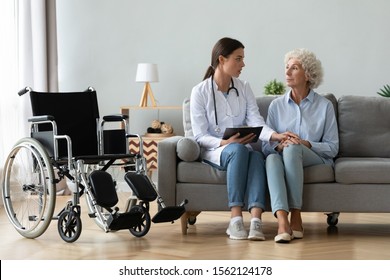 Serious Young Female Doctor Comforting Injured Disabled Handicapped Senior Lady Patient Tell Diagnosis Help Old Grandma With Disability Problem Talk At Medical Checkup Visit With Wheelchair At Home