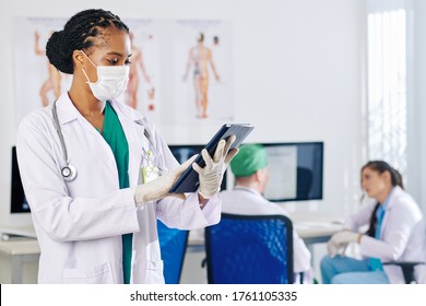 Serious young female general practitioner in medical mask reading medical history on digital table when standing in staff lounge
