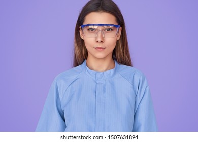 Serious Young Female In Lab Safety Goggles And Blue Coat Looking At Camera During Work Against Violet Background