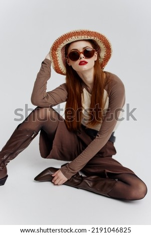 A serious young female model in a hat, glasses and boots is sitting on the floor in a white studio. Model catalog photography