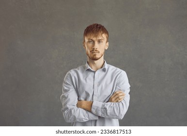 Serious young ginger guy in his 20s. Studio head shot portrait of man in his twenties with unsociable reserved introverted personality looking at camera standing arms folded against grey background