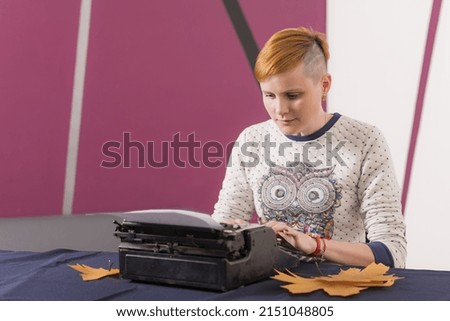 Serious young girl with red hair and stylish hairstyle sits at table and types text on old typewriter 