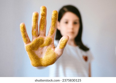 Serious young girl showing her palm hand covered of golden glitter while doing stop sign. Concept of harmful microplastics composition to humans and environment. Selective focus on hand.