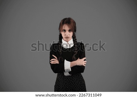 Serious young girl student in gothic black clothes with pigtails crossed arms on chest and looks at camera, isolated on gray wall studio background. Problems in study, stress, education and lifestyle