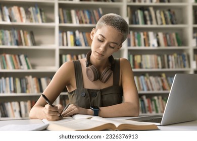 Serious young hipster student girl studying in university library, doing homework, writing notes, listening to webinar on laptop, working on research, essay, article at desk with open books - Powered by Shutterstock