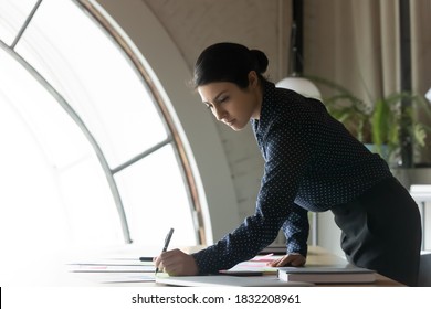 Serious young indian businesswoman busy working with financial company paperwork in office. Concentrated ethnic female employee brainstorm develop startup business project at workplace.