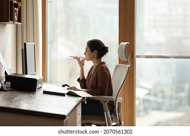 Serious young Indian businesswoman sit on chair at workplace holds smartphone talk to client on speaker phone, leaves voicemail, voice message communicates remotely use modern tech, connection concept