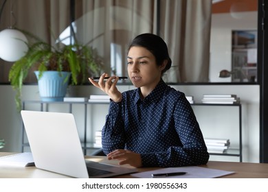 Serious Young Indian Businesswoman Work Online On Computer In Office Record Audio Voice Message On Smartphone. Focused Ethnic Female Employee Buys With Laptop Talk On Loudspeaker On Cellphone.