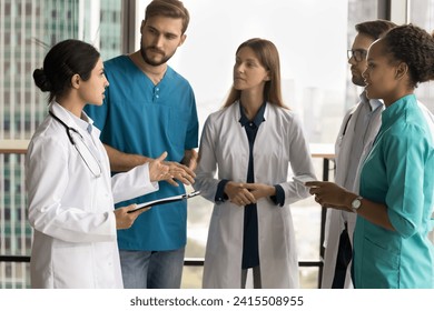 Serious young Indian doctor talking to medical diverse staff of city hospital, speaking to colleagues, practitioners in uniforms, standing in circle in office hall, discussing teamwork - Powered by Shutterstock