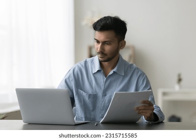 Serious young Indian entrepreneur man reviewing paper documents at work table, working at laptop, looking at screen, checking contract, agreement, consulting online legal service - Powered by Shutterstock