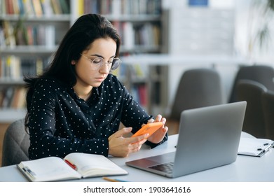 Serious young lady using smartphone, sit at the desk. Pretty woman in stylish wear browsing internet, chatting with colleagues or replies to an email - Shutterstock ID 1903641616