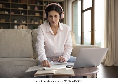 Serious young Latin student girl in big pink headphones studying at home, listening to audio lecture, workshop, reading learning textbook, open books, making summary, writing notes - Shutterstock ID 2139745003