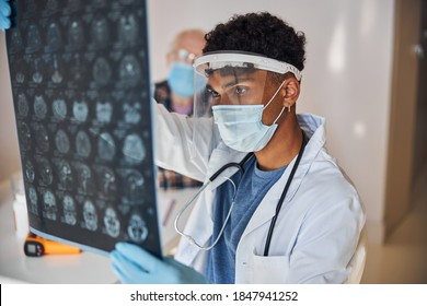 Serious Young Male Doctor In A Face Shield And A Mask Examining The MRI Scans