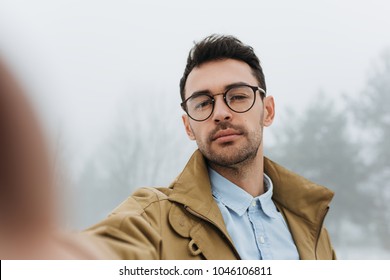 Serious Young Male Is Making Self Portrait On Camera Outdoors. Handsome Traveler Man Wear Coat, Blue Shirt And Eyeglasses Making Selfie And Smiling While Standing Against Grey Misty Nature Background.