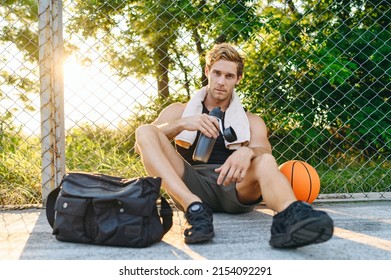 Serious Young Man 20s Wearing Sports Clothes Training Sit On Ground Bag Ball Drink Water Bottle Rest Look Camera Leaning On Metal Grid At Basketball Playground Court Outdoor Courtyard Sport Concept