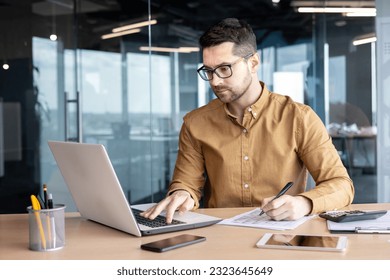 Serious young man accountant, auditor, analyst works in the office at the table behind the laptop and with documents. Writes invoices, makes analysis, fills out financial reporting and taxation.