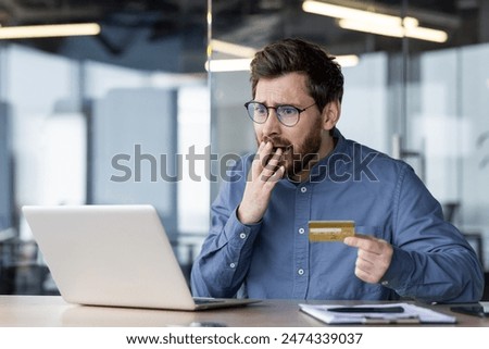 Serious young man businessman has financial problems with online account, cyber fraud. Sitting at the desk in the office, holding a credit card in his hand, looking shocked at the account screen.