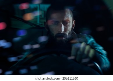 Serious Young Man Driving A Car At Night. Bokeh Lights Reflecting From The Windshield, Motion Blur