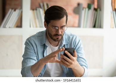 Serious young man in glasses look at cellphone screen read news online on gadget. Concentrated Caucasian male use smartphone consult client on web, browse internet on device. Technology concept. - Shutterstock ID 1916411651