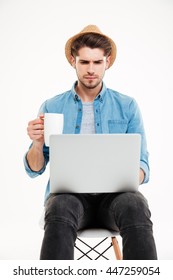 Serious Young Man In Hat Drinking Coffee And Using Laptop Over White Background