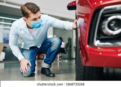 Serious Young Man In Medical Mask Checking Tires Of Car In Dealership