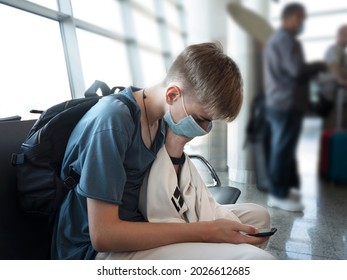 Serious Young Man Teenager 15-18 Years Old Typing A Message On The Phone While Sitting In The Airport Lounge. Young Student In Casual Clothes And A Protective Face Mask Using A Smartphone.