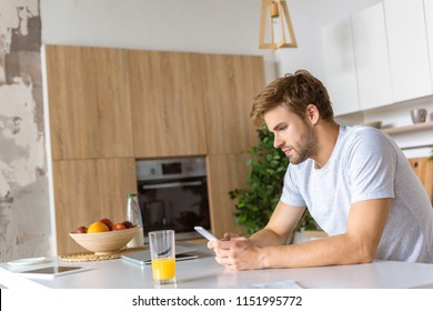 Serious Young Man Using Smartphone At Kitchen Table With Juice And Laptop 