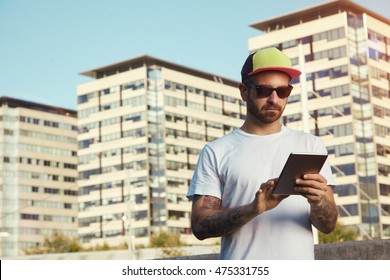 Serious Young Man Wearing White Plain T-shirt And Red, Yellow And Black Trucker Hat Looking At His Tablet Against City Buildings And Sky Background