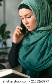 Serious Young Muslim Woman In A Hijab Is Talking On Mobile Phone With A Notebook In Her Hands While Sitting At A Table In A Home Interior.Business,freelance,diverse People Concept.