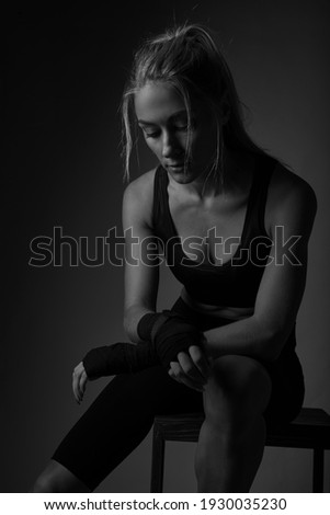 Serious young professional female kickboxer wearing black sportswear and bandages on hands, posing in dark atmosphere. Fighter having rest after training