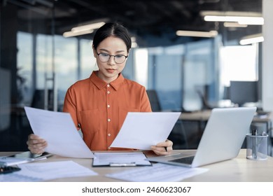 A serious young professional woman in a stylish orange shirt reviews paperwork intently, surrounded by laptops in a well-lit office setting. Her focus and determination are evident. - Powered by Shutterstock