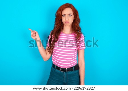 Serious young redhead woman wearing pink striped T-shirt over blue background smirks face points away on copy space shows something unpleasant. Look at this advertisement. Big price concept